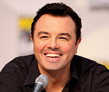 A man with short black hair and a black shirt in front of a microphone, and he is laughing.