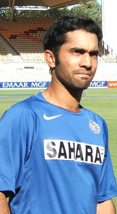 Brown-skinned young man, not clean shaven, wears a sky blue shirt with the words "SAHARA" on it stares forward. In the background is lawn of a sporting ground and a grandstand.