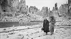 A soldier holding the arm of an elderly lady in a debris-strewn street, with ruined buildings in the background