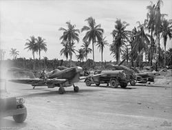 Black-and-white photo of two single-engined monoplane aircraft and a fuel tanker on a dirt clearing. Tropical palms are visible in the background.