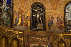 A stained glass window shows the crucifixion. On either side mosaics show angels holding symbols of the Passion and Glory of Christ. Beneath is a mosaic depicting Christ and His apostles celebrating the Passover.
