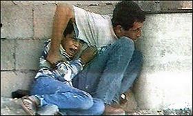 A man with black hair wearing blue jeans and a white t-shirt crouches behind a wall and a white concrete cylinder. With his right hand, he is grasping the arm of a young boy, also with black hair, who is crouching on the ground behind him. The boy is wearing blue jeans, brown sandals, and a blue and white top. His right hand is holding onto the man's t-shirt. He looks as though he is crying. Behind them, the wall is made up of concrete blocks. The man's head is slightly down, and he is looking to his left.