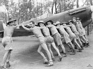 Nine men wearing shorts, seven of them wearing wide brimmed hats, push a propeller-driven aircraft.