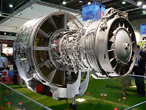 An exposed jet engine at a trade show. The rear of the polished metal fan case is visible on the left. The outer casing of the compressor section, covered in fuel lines and electrical wires is in to the right of the fan case. The right of the image shows the back of the engine, the exhaust area of the turbine section.