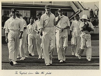 A group of ten men are walking onto a grass playing field in front of a building which has spectators sitting in front. The men are all dressed in white shirts and trousers while most of them are wearing cricket caps. One man has pads and wicketkeeping gloves on.