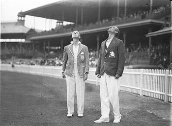 Two men are standing on a cricket field looking in the air. Both are wearing dark blazers and white trousers. There is a crowd in the background behind a white fence.