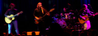 Four members of The Church are performing on-stage. Koppes is facing forward and strums his guitar. Kilbey is playing a bass guitar and singing into a microphone. Powles is set back, obscured by his drum kit. Willson-Piper is partly turned to his left and is strumming a guitar.