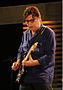 "A colour photo of Robbie Robertson on a stage. He is wearing a purple shirt while playing a six string fender guitar."