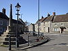 Street scene. Stone cross on a pillar rising from 5 step plinth. Iron lampost left and right of the road are stone terraced houses.