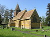 Yellow stone building with red tiled roof and square tower with short spire. Foreground is grass with gravestones.