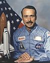 man with mustache in astronaut uniform, model of space shuttle to side, U.S. flag in background