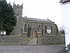 Gray bstone building with square tower to the left hand end, partially obscured by tree. In the foreground is a stone wall separating the church from the road.