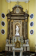 A white statue of a saint, pierced by golden arrows, is framed by marble. In the foreground, inside a perspex case, is a gold reliquary, with a statue of a man seated on a throne, holding an orb in his left hand, surrounded by a golden frame.