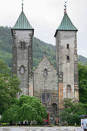 Stone  Romanesque church with two  towers and a  lower, pitched roof entrance hall between them. The towers have slightly curved  bronze roofs.