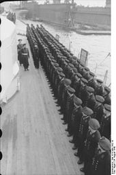 Two men walking by a row of men on the side of ship.