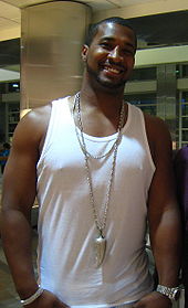 A smiling black man with very short, dark hair, wearing a white sleeveless shirt.