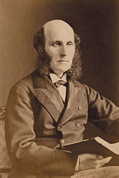 A well dressed middle aged man with a bald head and bushy sideburns, wearing a dark suit and carrying a large book