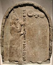 Stone stele with a carving depicting a man with a beard, carrying a tall staff and wearing a robe and conical hat, gesturing to three symbols representing the moon, sun and Venus.
