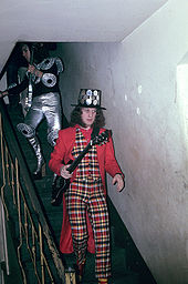 Two men walking down some stairs. In front, the man is wearing a suit, and a jacket on top. He carries a guitar. His hair comes down to his shoulders, and he has large sideburns. On his head is a top hat, covered with large coins. The man following him is wearing metallic plates on his knees, arms and shoulders, and is wearing platform shoes. He carries a guitar, and on his arms is some jewellery. He is wearing a hood of some sort on his head.