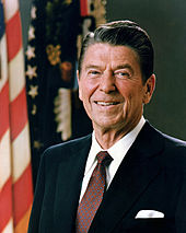 A photograph of a dark-haired, old man, wearing a suit and smiling in front of an American flag