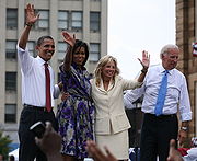 Barack and Michelle Obama and a woman and a man on an outdoor stage. The first three smile and wave. The men wear suit pants, white shirts with the sleeves rolled up, and ties. Michelle is in a colorful print dress and the other woman is in a creme business suit.