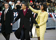 Barack and Michelle Obama hold hands and smile while walking; she waves to a crowd. She wears a gold embroidered dress and coat; he wears a black overcoat and burgundy scarf. A serious man in a dark suit watches nearby.