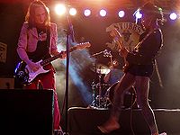 Three musicians performing on stage, man at left foreground is playing guitar, man in middle background on drum kit and woman at right foreground is playing guitar.