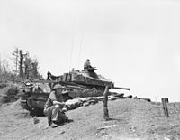 A main battle tank in a hull down position, its turret traversed to aft. In the foreground is a soldier kneeling.
