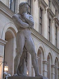 A marble statue of Spartacus in front of a neoclassical building. He is standing naked with his legs astride, looking into the middle distance with his left hand clenched against his chin, his right arm crossing his chest on which his left arm rests.