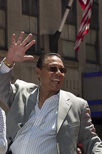 A man in a grey jacket and sunglasses wearing a white shirt with blue stripes waves.