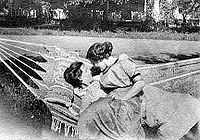Black and white photo of two women sitting in a hammock in turn of the 20th century dresses; one reclines and the other sits on her lap and wraps her arm around the other, both staring at each other
