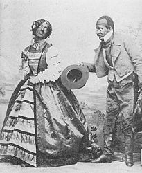 Two blackface performers on stage, one a man dressed in fancy woman's clothes, the other in dress attire, bowing with his hat in his hand.