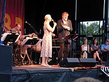 Full length portrait of young woman wearing a sleeveless dress on left, blond hair pulled back into ponytail, and middle aged male wearing a brown tuxedo on right, in brown longish hair, singing into microphones. Three people seated at right listening, guitar player visible in band at left.