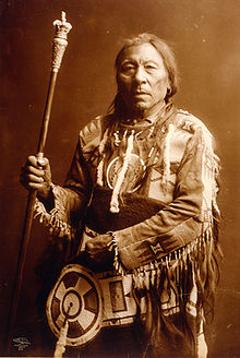 A sepia photograph of Aatsista-Mahkan (Running Rabbit). He is wearing what is usually described as a buckskin outfit. It is elaborate and he is holding a pole.