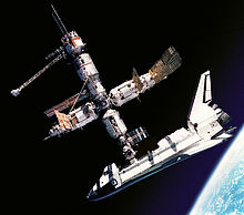 A cluster of cylindrical modules with projecting feathery solar arrays and a space shuttle docked to the lower module. In the background is the blackness of space, and, in the lower right corner, Earth.