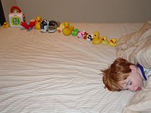 Young boy asleep on a bed, facing the camera, with only the head visible and the body off-camera. On the bed behind the boy's head is a dozen or so toys carefully arranged in a line, ordered by size.