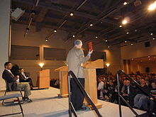 Man in his fifties standing and speaking wearing a tan suitcoat holding up a red book, man in his forties seated, large audience visible at right