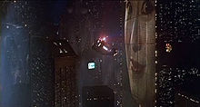 A screenshot of a police spinner flying through a cityscape next to a large building which has a huge face projected onto it. In the distance a screen can be seen with writing and pictures on it