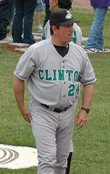 A man wearing a gray baseball uniform with "Clinton" written in green letters across the chest and a black cap on a baseball field