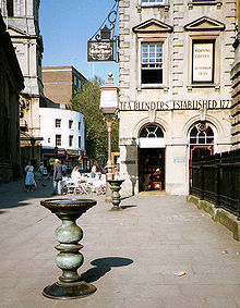 Two ornate metal pillars with large dishes on top in a paved street, with a eighteenth century stone building behind upon which can be seen the words "Tea Blenders Estabklishec 177-". People sitting at cafe style tables outside. On the right iron railings.
