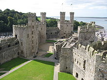 In a picture taken from a high vantage point is seen the courtyard of a castle, surrounded by walls and towers. In the background are seen wooded hills to the left, and ocean and shoreline to the right.