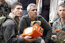 Three men, one of whom is holding a partially mangled red metal box