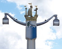 Post holding two lamps at the ends of two arms. The top of the post is decorated by a galleon; underneath the boat is a shield containing a white left and a blue right separated by a jagged line, on top of which rests a white bird carrying an olive branch.