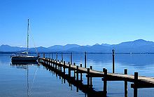Image of a pier extending out in to a lake, with a clear sky above and mountains in the distance.
