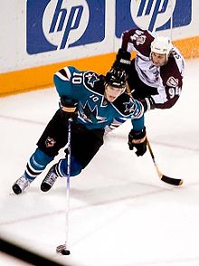 An ice hockey player dressed in a teal and black jersey cradling the puck with his stick in mid-stride. An opposing player is in pursuit behind him.