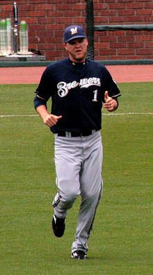 A man wearing gray pants, a navy blue jersey with "Brewers" written in white, and a navy blue cap bearing a white "M" running on a baseball field