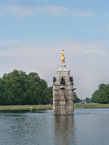 a golden female statue on top of a white marble column, smaller black statues adorn the column and there is a water jet into a surrounding pool. In the background are trees