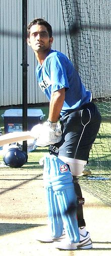 Brown-skinned young man, not clean shaven, wears a sky blue shirt with the words "SAHARA". He is wearing white sports shoes, navy shorts and black stockings, is wearing light blue cricket pads on his legs, white gloves, is holding a bat and is capless, in batting stance on a cricket pitch.