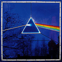 A small bright white equilateral triangle stands slightly above the centre of a dark blue image, framed by silhouetted trees and assorted stars.  At each point of the triangle a straight black line heads to the edge of the image, which has a grey and blue border.  The two lower black lines from the points of the triangle are linked by a horizontal black line.  A perfectly straight bright white line enters from the middle left edge of the image, angled slightly upward to meet the left side of the triangle.  Inside the triangle the white line expands slightly, fading to blue as it reaches the centre.  On the right side of the triangle a thick bar composed of red, orange, yellow, green, blue, and violet angles downward to the middle right edge of the image.  Small black lines separate the colours, and smaller black lines split each colour into sections.