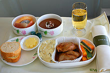 Business class meal. Cloth-covered tray with napkin, tall glass, two round bowls with fruits and chocolate cake; plate with bread and butter, large dish with potatoes, meat, and vegetables.
